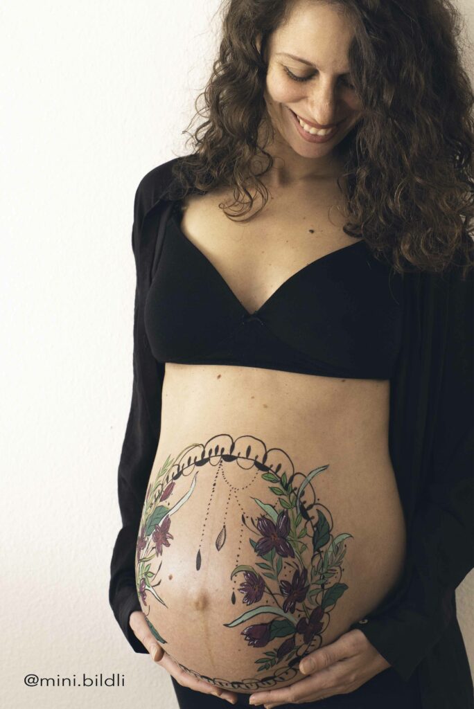 Pregnant woman with belly painting of flowers and mandala smiling while holding her belly with her hands and looking at her.