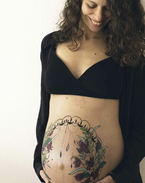 Pregnant woman with belly painting of flowers and mandala smiling while holding her belly with her hands and looking at her.